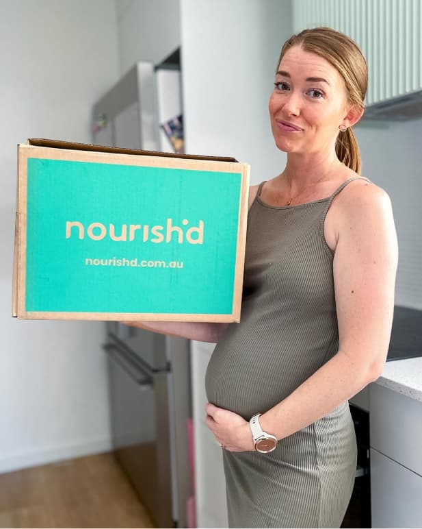 An expecting mum holding a Nourish'd box with one hand and her stomach with the other.