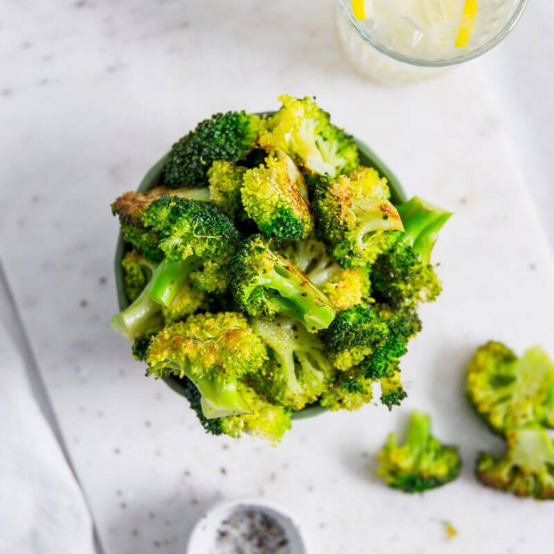 Bowl of roasted broccoli florets on a white surface with a glass of lemon water and scattered broccoli pieces nearby.