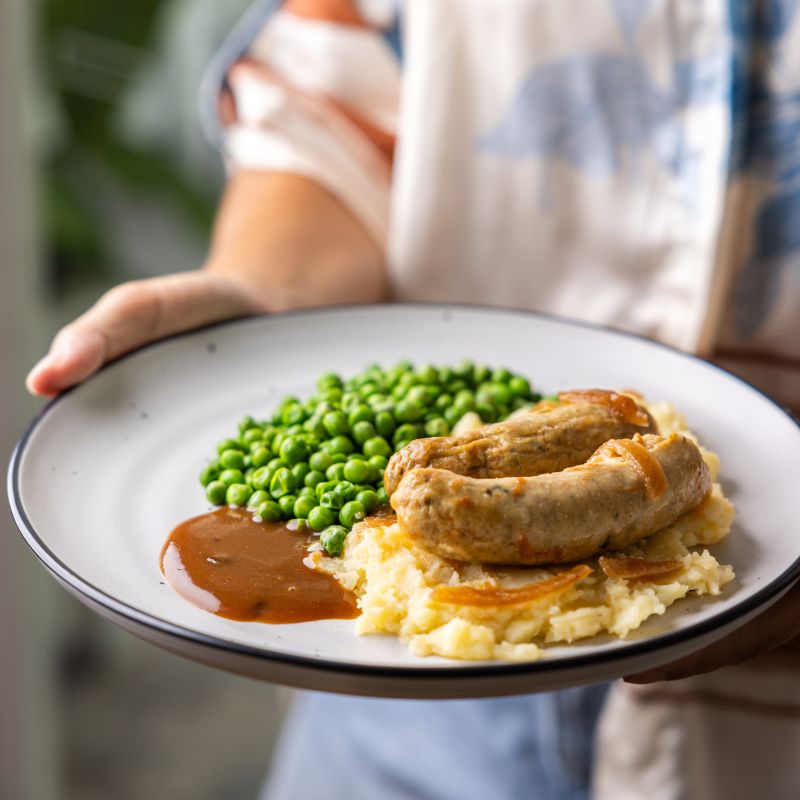  A side-angle shot of a woman holding a plate of Bangers and Mash. She is wearing a silky cream and blue top, adding an elegant touch to the homestyle dish.