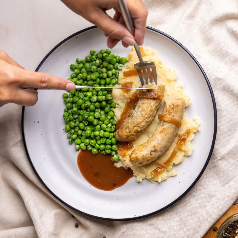 An action shot of someone cutting into the sausage with a fork and knife, revealing the juicy texture. The hearty mash and gravy create a mouthwatering scene.