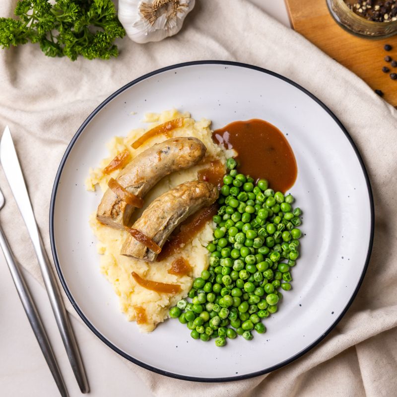 An action shot of someone cutting into the sausage with a fork and knife, revealing the juicy texture. The hearty mash and gravy create a mouthwatering scene.