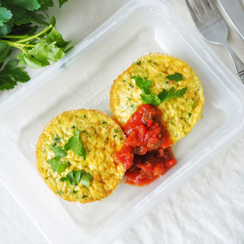 A top-down photograph of two muffin shaped frittata's in a serving container, sprinkled with some green herbs and finished with a dollop of tomato salsa. Beside the container lays a knife and fork and some extra herbs.
