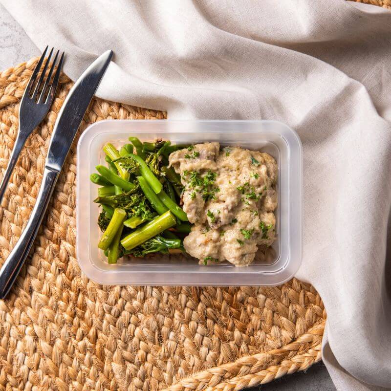 Clear container shot of the Honey Mustard Chicken, beautifully arranged to show off the creamy mustard sauce and spring onion garnish. The background includes the rattan placemat and grey marble countertop for a clean, cohesive look.