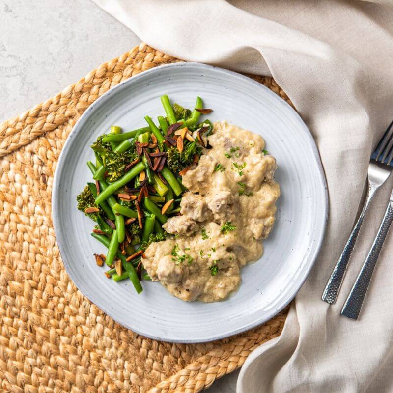 Flat lay image of Honey Mustard Chicken served on a blue-hued plate. The plate is placed on a rattan placemat resting on a light grey marble countertop. To the right of the plate is a beige cloth with a knife and fork placed neatly on top. The dish is garnished with freshly chopped spring onion, adding a pop of green