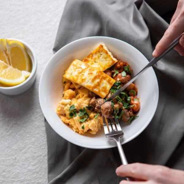 An action shot of the Breakky Bowl with two hands holding a fork and knife, cutting into a sautéed mushroom. The bowl sits on a neutral surface, with a white ramekin of sliced lemon visible in the background