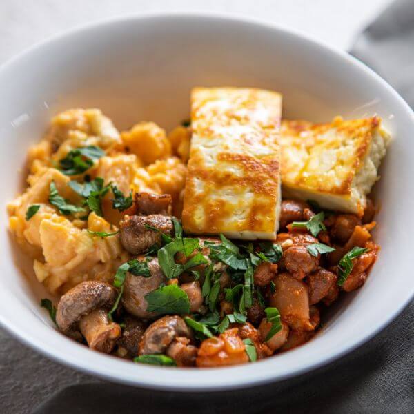 A close-up view of the Breakky Bowl, highlighting the fluffy scrambled eggs, golden grilled haloumi, rich smoky baked beans, and sautéed mushrooms. Chopped spring onion is sprinkled on top for a fresh garnish.