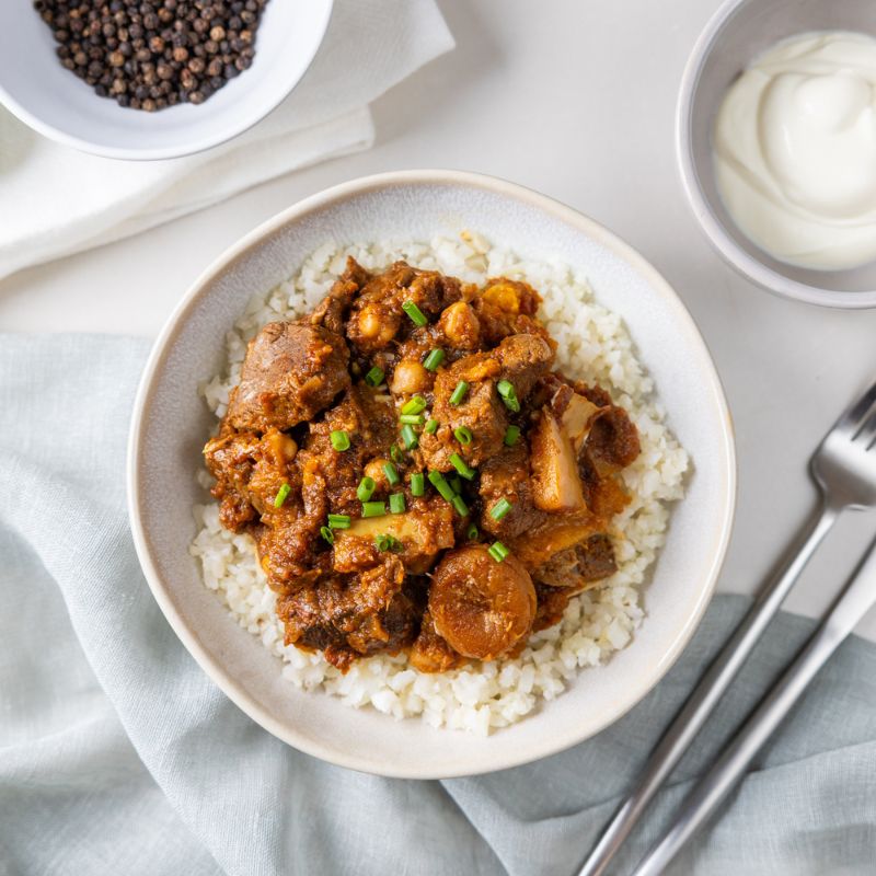 A top-down view of Moroccan Beef and Apricot Tagine with Cauliflower Rice served in a white bowl on a white countertop. The dish features tender beef, dried apricots, and pumpkin on a bed of cauliflower rice, sprinkled with exotic spices. A light grey cloth is tucked under the bowl. A fork and knife sit to the right, while a ramekin of Greek yogurt is in the top right corner and a ramekin of cracked peppercorns in the left corner