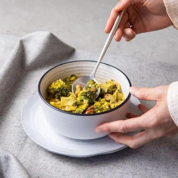 Action shot of two hands, one holding the bowl of Thai Green Curry and the other spooning into the dish, highlighting the rich, colorful curry packed with vegetables.