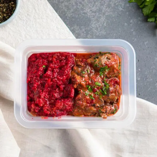 A clear plastic container of Beef Stroganoff, showcasing the creamy beef and vibrant beetroot and sweet potato mash. Chopped spring onions sit on top as a garnish. The container rests on a white cloth, with a ramekin of cracked peppercorns visible nearby for added flavor.