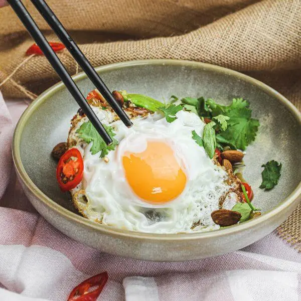 A side angle photograph of the fried egg, a top a bowl of nasi goreng. A hand is holding a pair of chopsticks, as if about to use them to eat. The bowl is nestled on two different types of cloth that are decorated with chopped chilli.