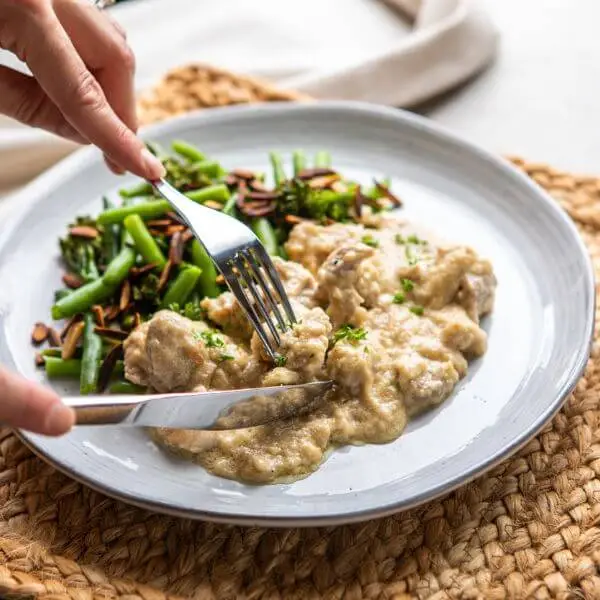 Action shot of two hands using a fork and knife to cut into the Honey Mustard Chicken. The same blue-hued plate, rattan placemat, and light grey marble countertop elements create a cohesive and inviting scene.
