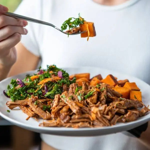 A photo of a person holding a white plate, filled with pulled barbeque brisket, roast sweet potato and kale slaw. Another hand is holding a fork with bits of each food component on it, as if about to eat.